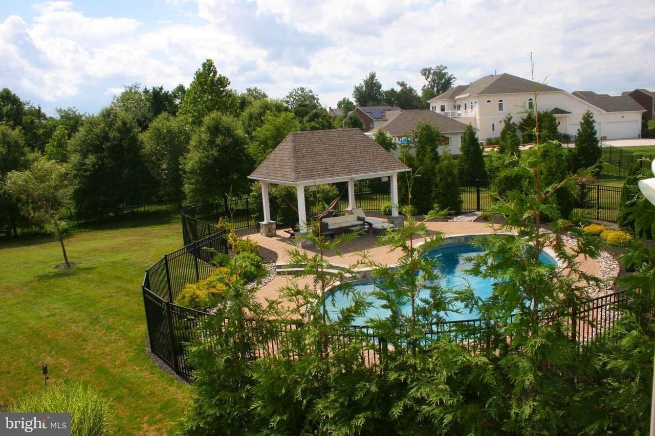 A high-angle shot of a large house with a fenced backyard oasis with trees, a covered patio, and blue swimming pool